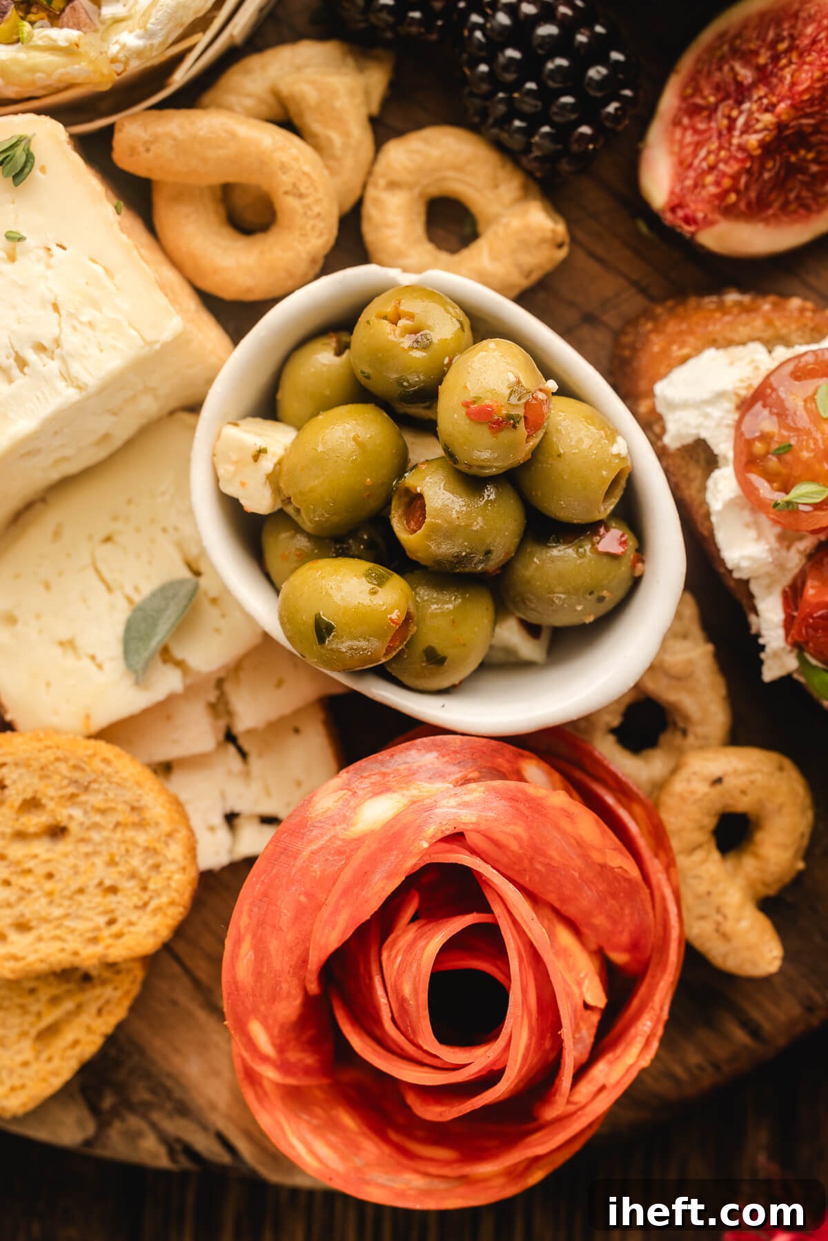 Overhead close-up of a salami rose on a charcuterie grazing table with olives, crackers, cheese, and fruit. 