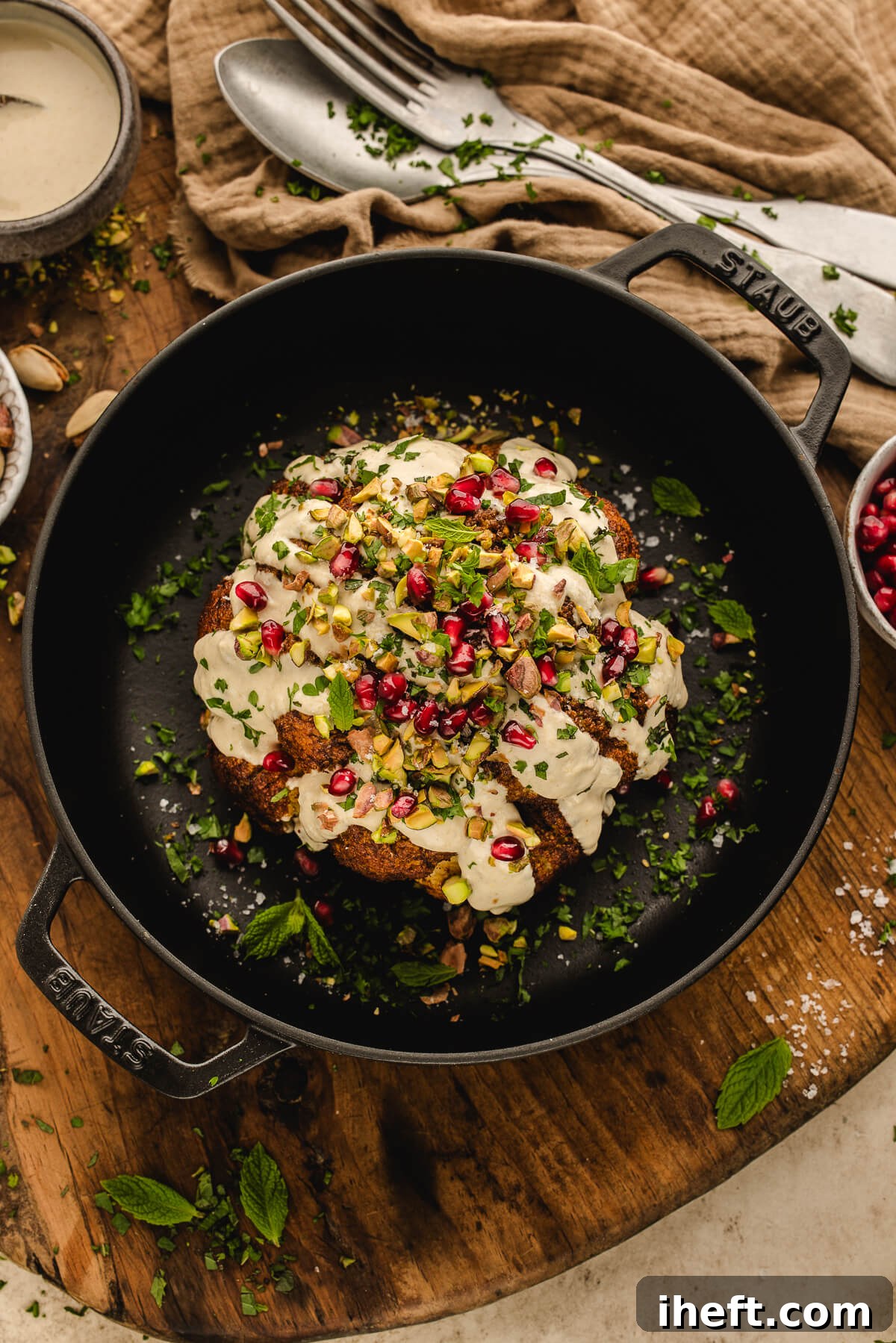 Whole roasted cauliflower served in a Staub cast-iron skillet topped with creamy tahini sauce, chopped pistachios, pomegranate seeds, and fresh herbs.