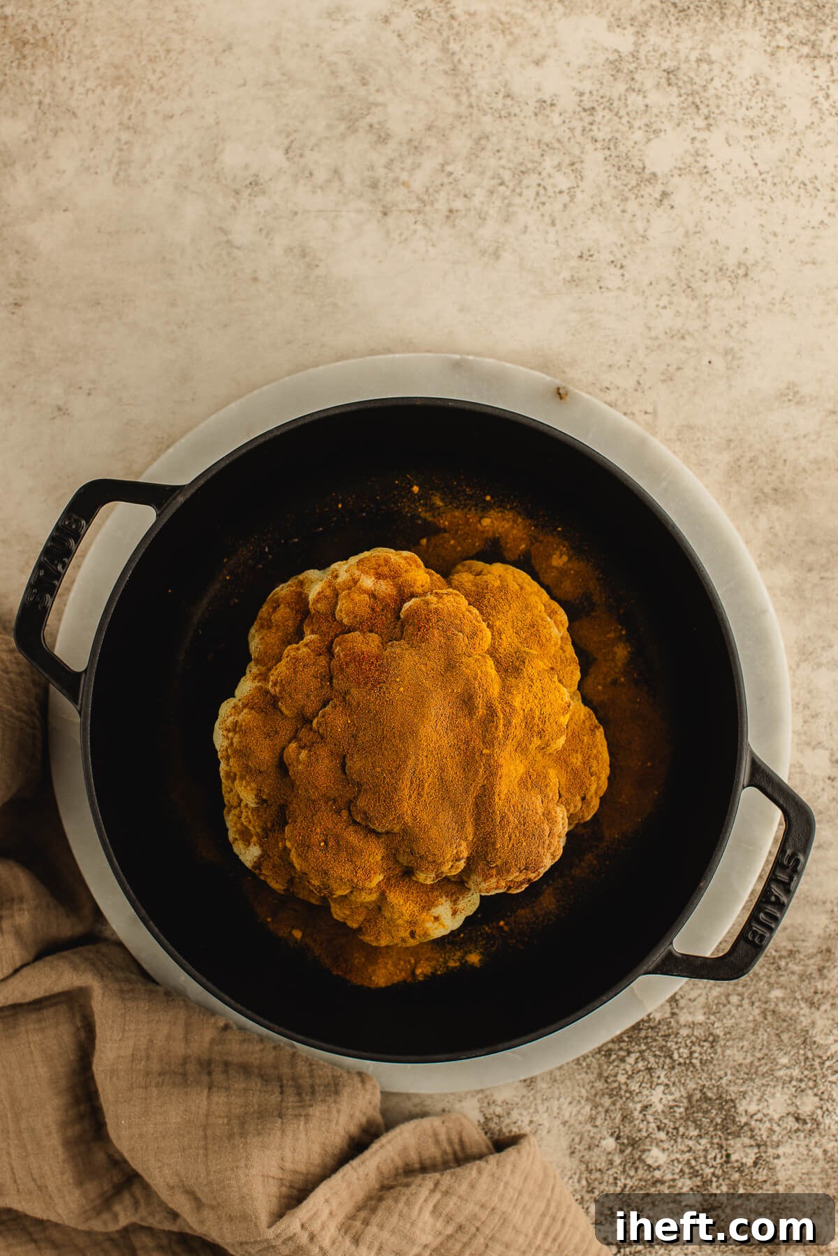 A whole cauliflower generously covered in warm spices and olive oil in a black cast iron pot, prepared for roasting.