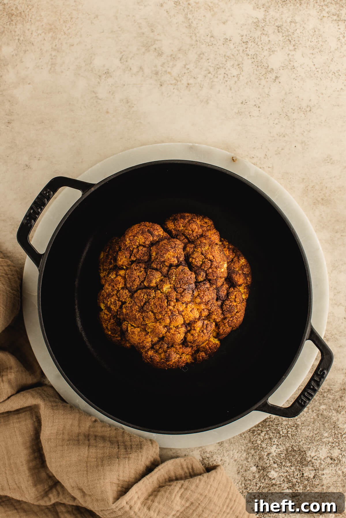 An overhead view of a perfectly spiced and golden whole roasted cauliflower in a black cast iron pot, fresh from the oven.