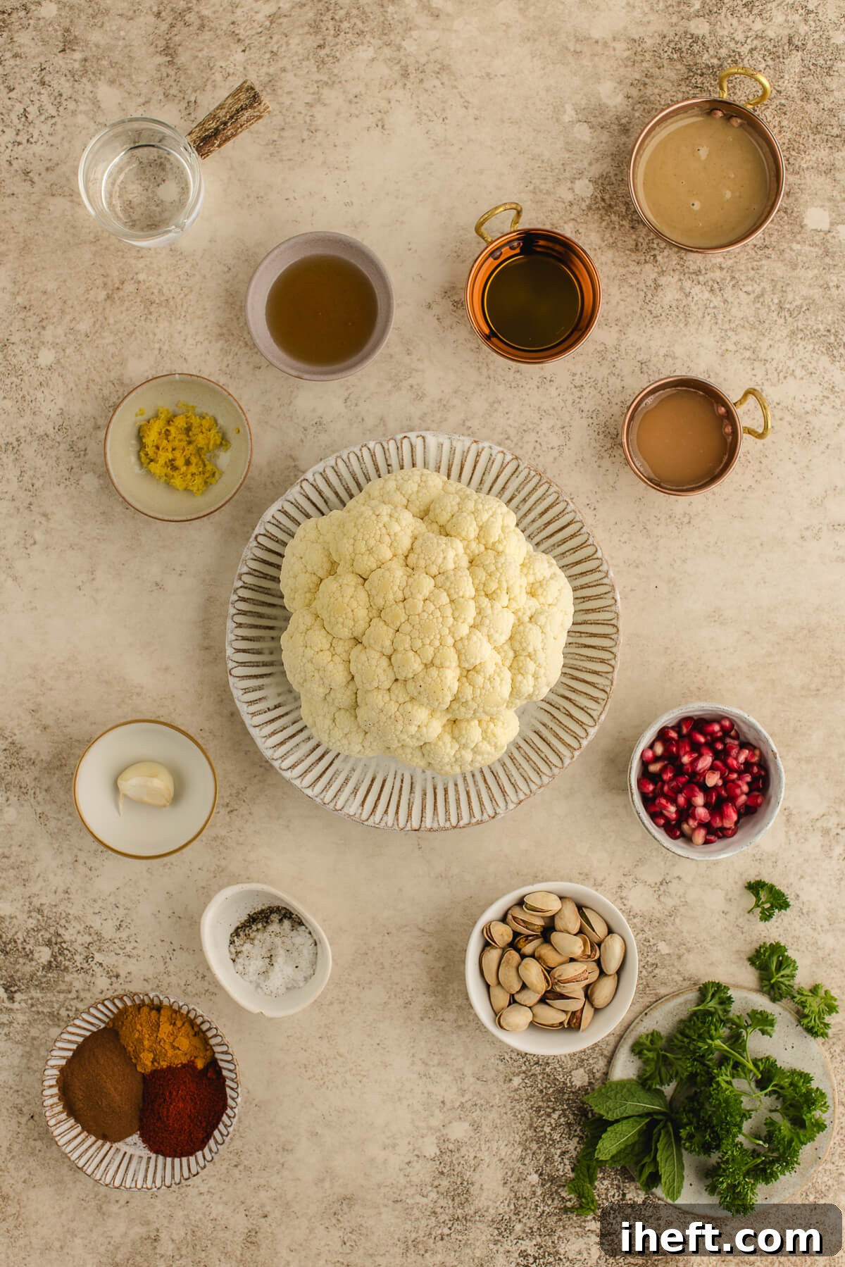 Ingredients for whole roasted cauliflower including fresh cauliflower, tahini, olive oil, lemon zest, garlic, herbs, pistachios, and pomegranate seeds arranged on a neutral surface.