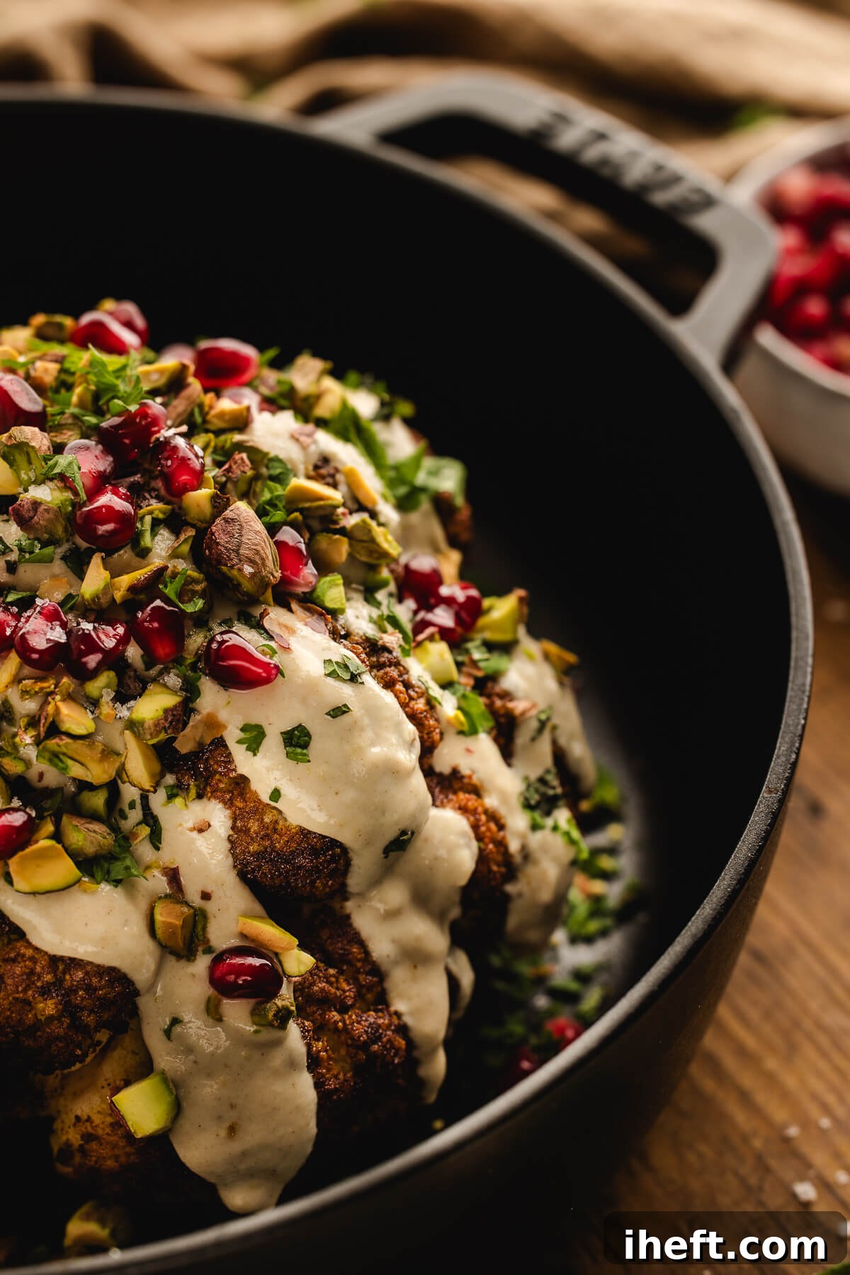 Whole roasted cauliflower in a cast iron skillet, topped with creamy tahini sauce, chopped pistachios, pomegranate seeds, and fresh herbs.