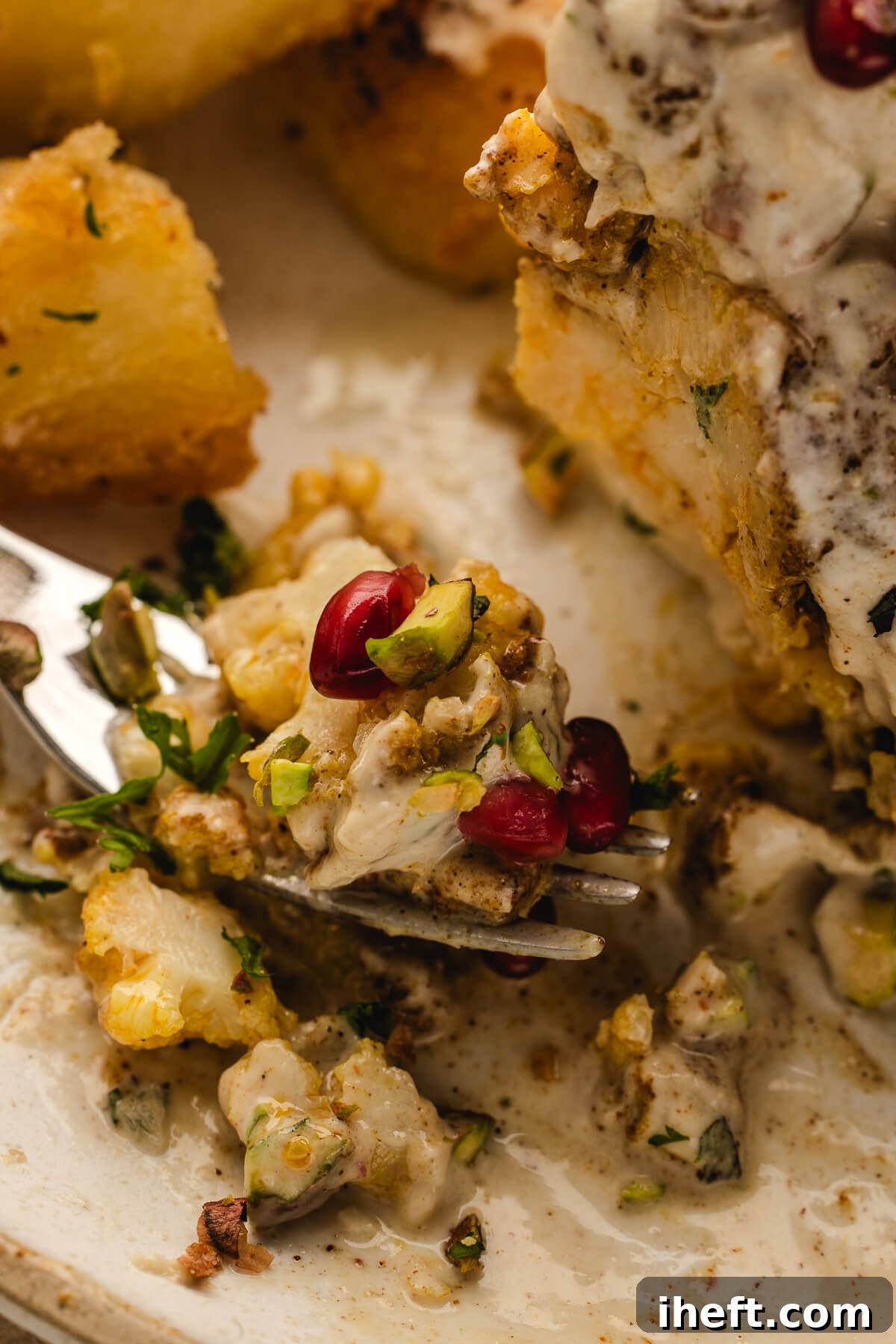 Close-up bite of roasted cauliflower with creamy tahini sauce, pistachios, and pomegranate seeds on a fork.