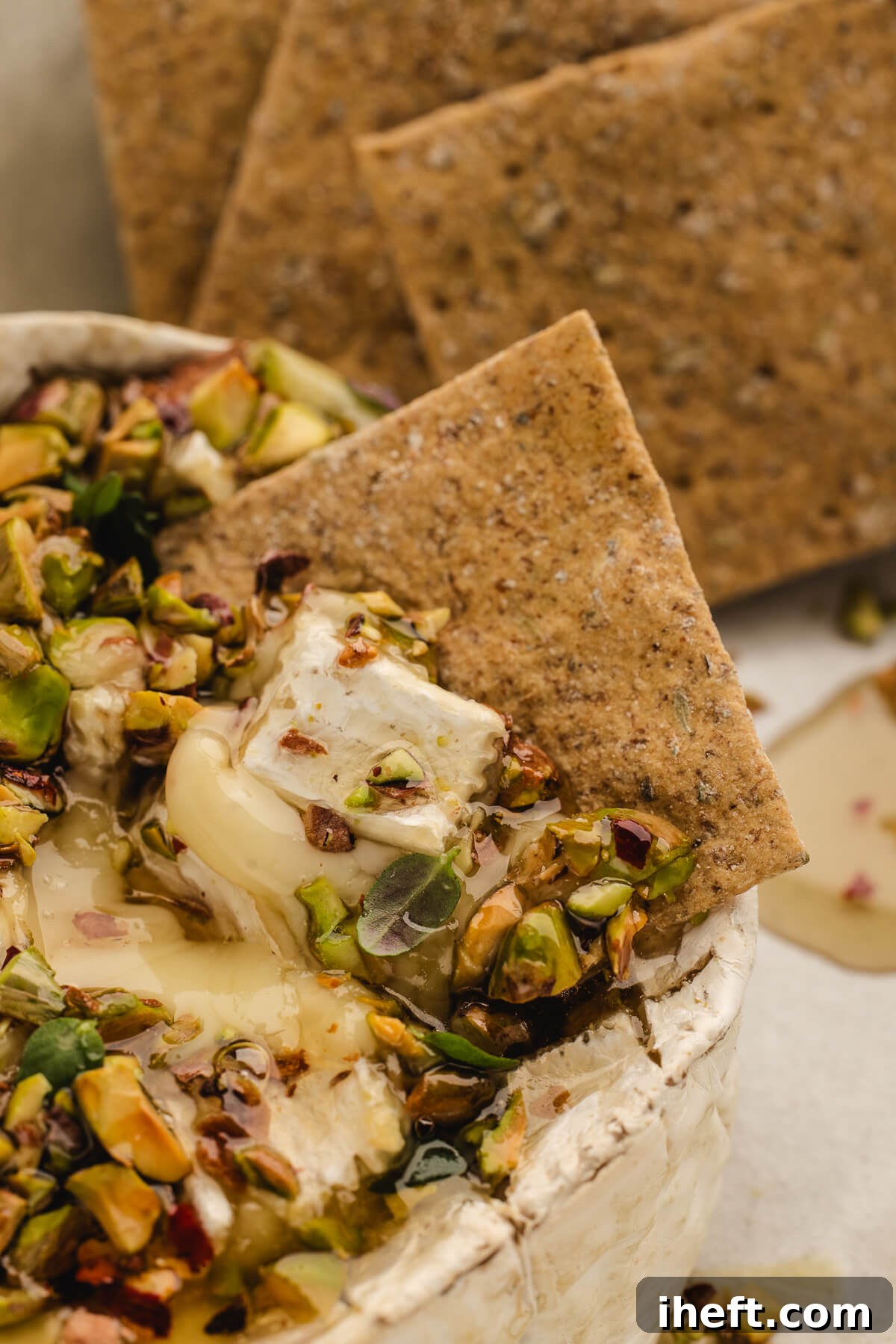 Melty baked Brie with chopped pistachios and hot honey surrounded by rustic crackers on a serving board.