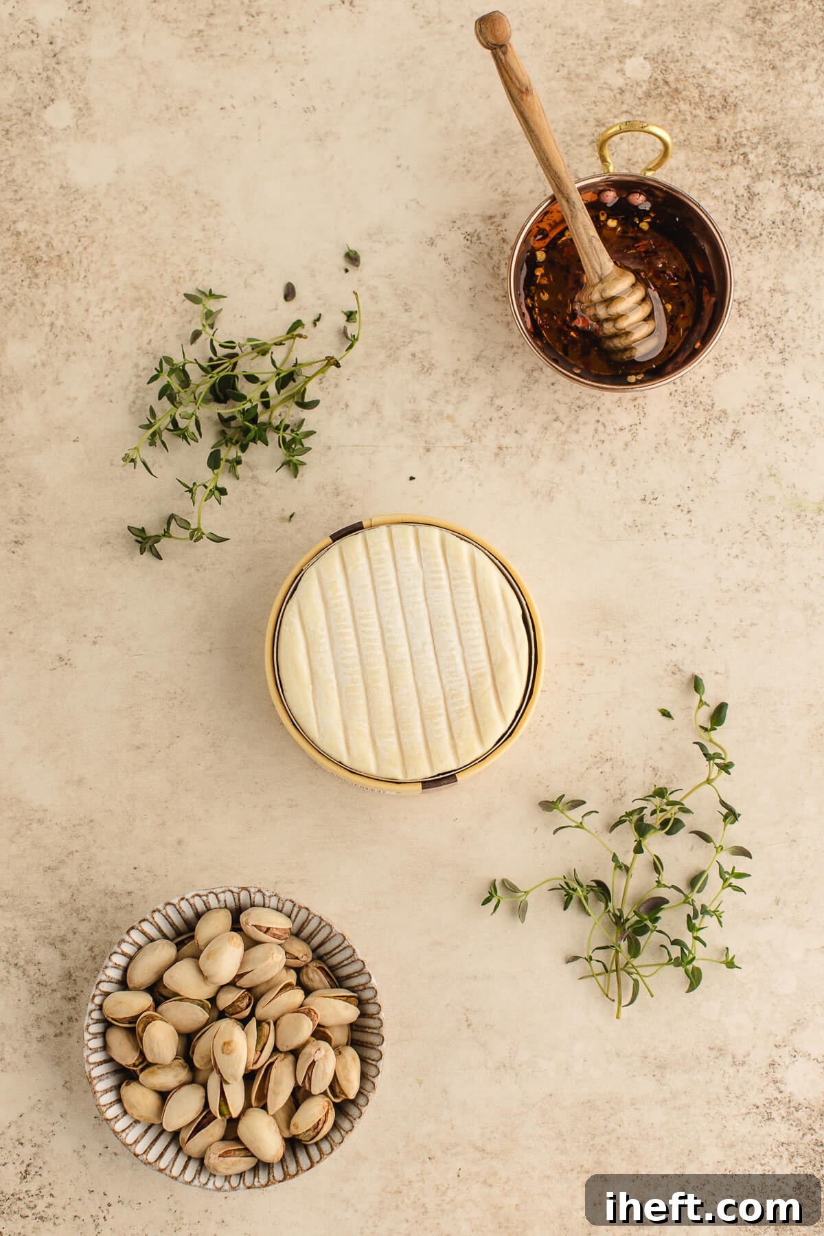 Ingredients for hot honey baked Brie including a wheel of Brie, pistachios, thyme, and hot honey arranged on a neutral background, ready for preparation.
