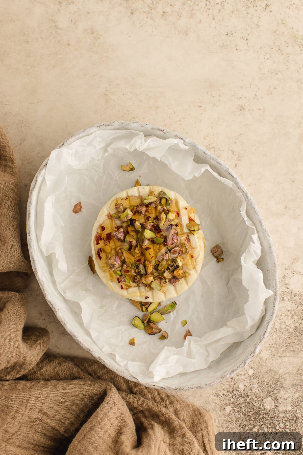 Unbaked wheel of Brie scored and topped with pistachios and thyme on parchment, ready for a holiday appetizer, showcasing the prepared cheese.
