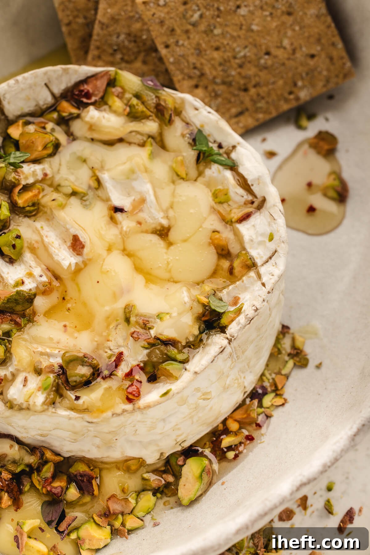 Close-up of melty baked Brie topped with chopped pistachios and herbs with crackers on the side, highlighting the gooey texture.