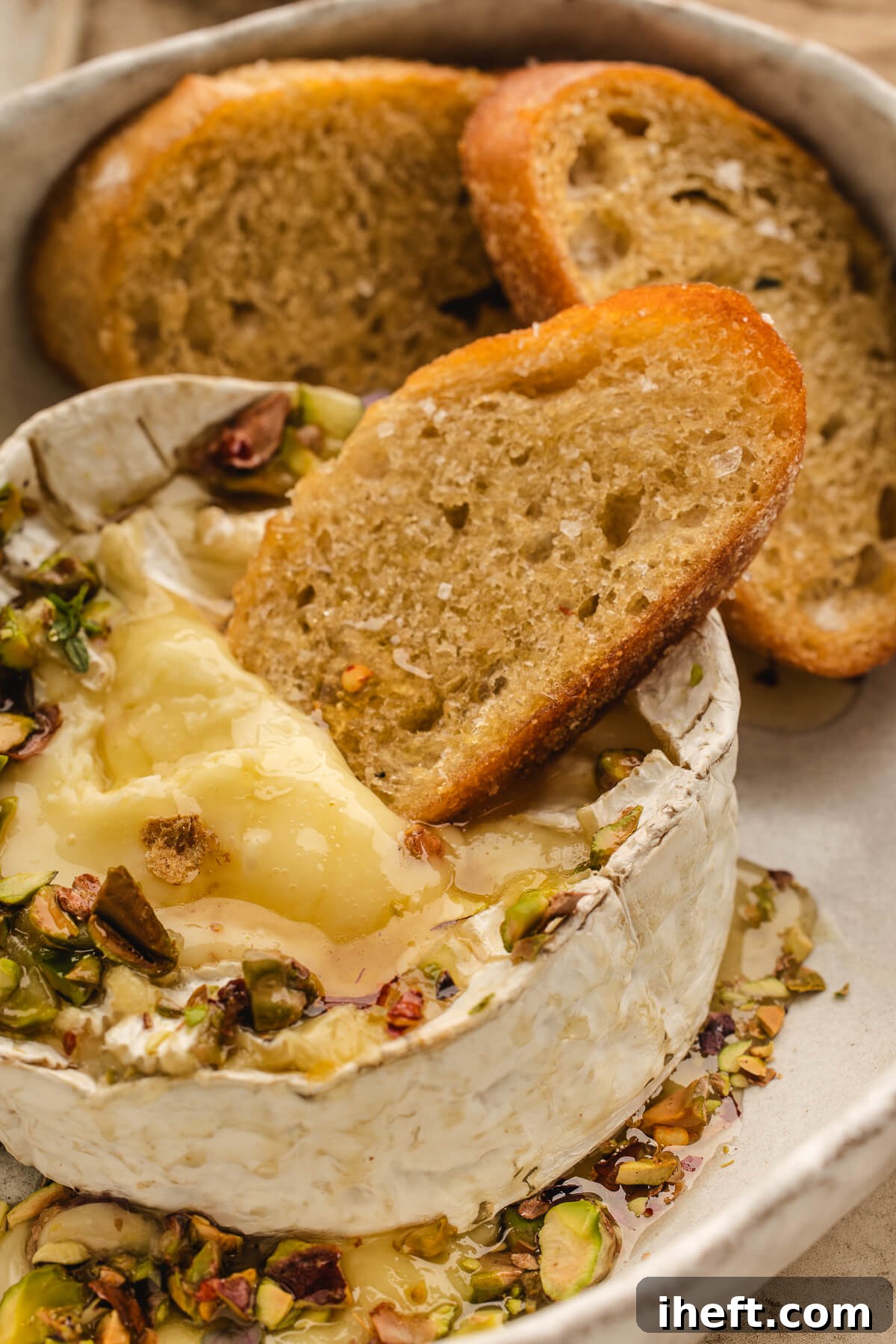 Close-up of warm baked Brie topped with chopped pistachios, served with toasted crostini, showcasing its inviting presentation.