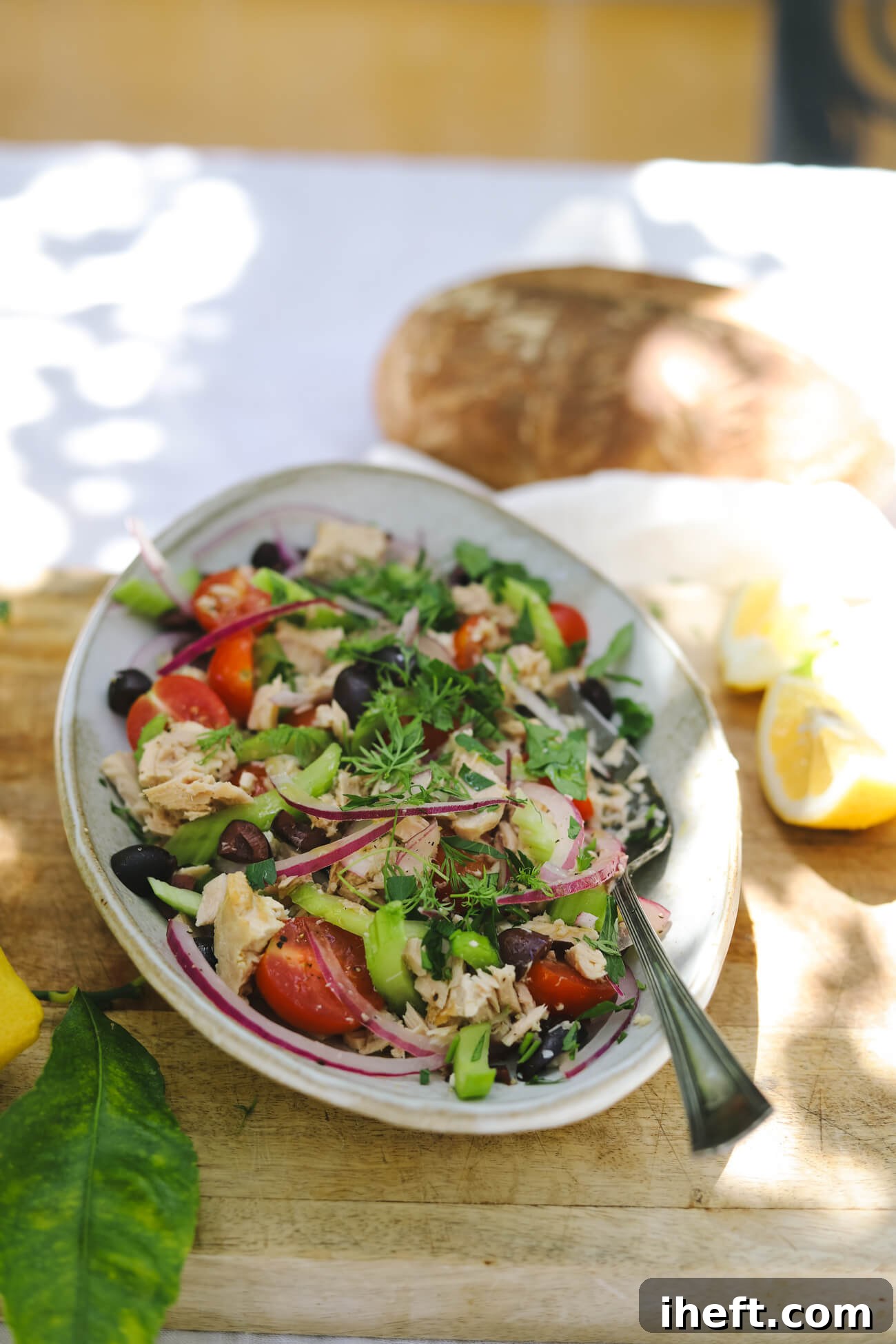 Mediterranean tuna salad in a bowl with albacore tuna, cherry tomatoes, kalamata olives, celery, red onion, parsley, lemon, and crusty bread. The bowl sits on a sunny outdoor table.