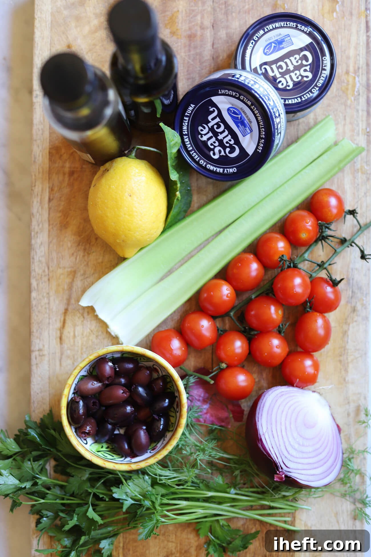 Mediterranean Tuna Salad Ingredients Ingredients for Mediterranean tuna salad including albacore tuna, cherry tomatoes, celery, red onion, kalamata olives, parsley, lemon, olive oil, and red wine vinegar on a wooden board.